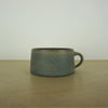 handmade ceramic mug by aku ceramics. simple shape in a blue green glaze. on a wooden table with a white wall. 