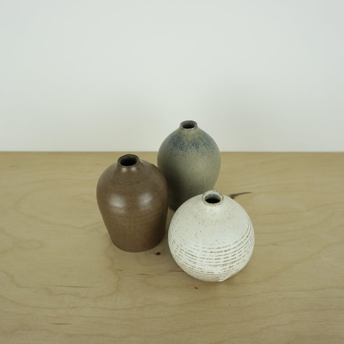 three mini handmade ceramic vases by Aku ceramics on a table. one white, one blue and one brown. 