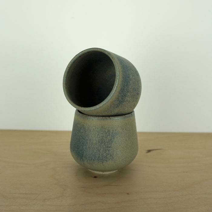 small handmade blue green cup, sitting on an angle inside another cup on a wooden table and a white background. 