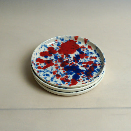 Stack of round ceramic plates with red and blue splatter design on a plain background. Splatter Saucer | Anna Jones Ceramics | Shop Fodder
