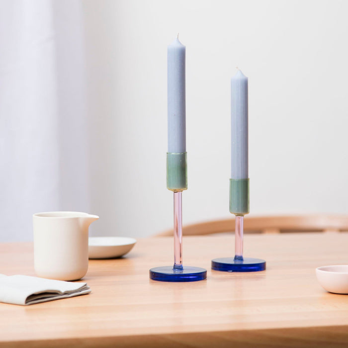 two glass candlestick holders on a table with a jug and salt dish 