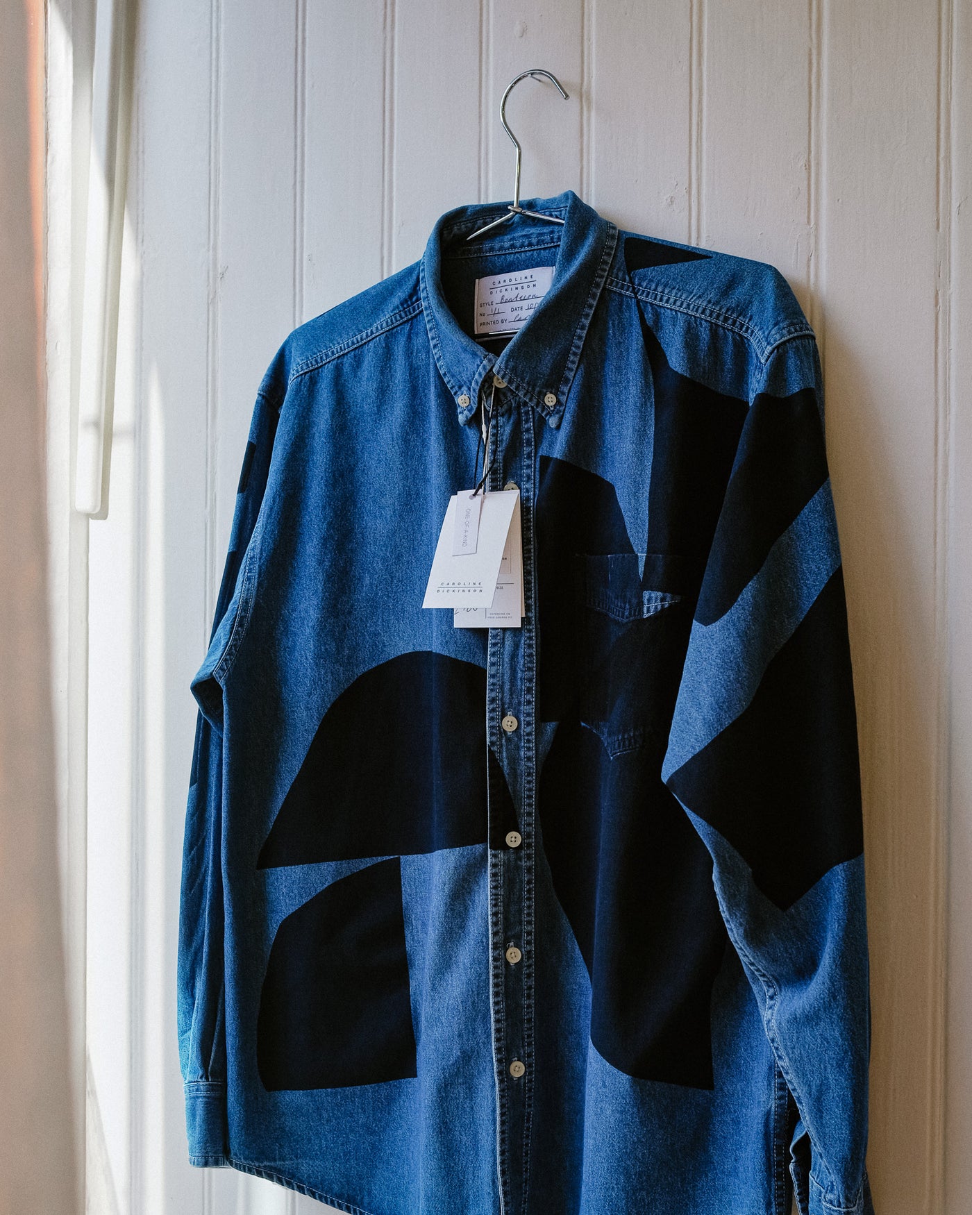 Blue denim shirt with black geometric patterns hanging on a white wall. Caroline Dickinson | Shop Fodder