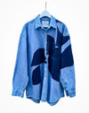 A blue denim button-up shirt with abstract dark blue patchwork panels, hanging on a white hanger against a plain white background. Caroline Dickinson Shirt | Fodder