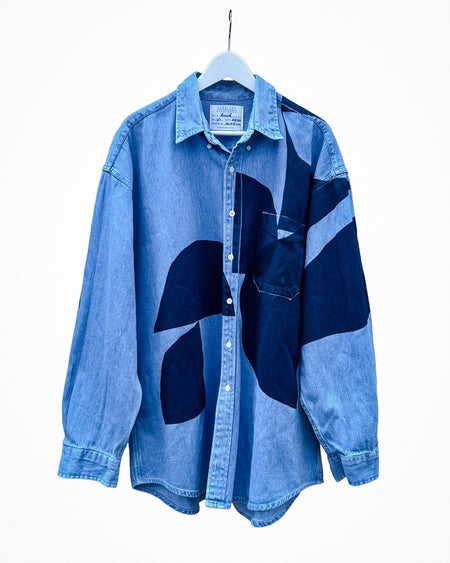 A blue denim button-up shirt with abstract dark blue patchwork panels, hanging on a white hanger against a plain white background. Caroline Dickinson Shirt | Fodder