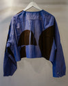 A cropped blue long-sleeve shirt with large abstract brown color-block panels on the back, hanging on a white hanger against a plain light gray wall. Caroline Dickinson Shirt | Fodder