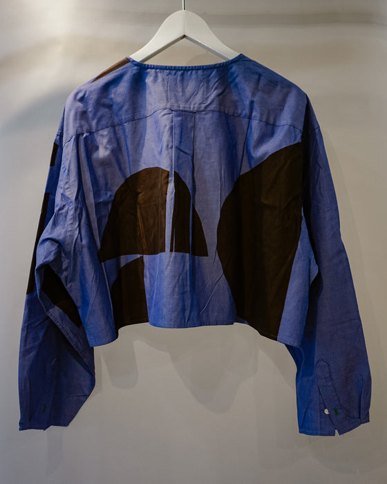 A cropped blue long-sleeve shirt with large abstract brown color-block panels on the back, hanging on a white hanger against a plain light gray wall. Caroline Dickinson Shirt | Fodder
