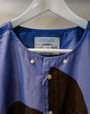 A close-up of a Caroline Dickinson shirt in blue and brown, hanging on a white hanger. The shirt features a textured blue body, a brown color-blocked panel on the lower right, and white buttons. Caroline Dickinson Shirt | Fodder