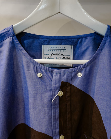 A close-up of a Caroline Dickinson shirt in blue and brown, hanging on a white hanger. The shirt features a textured blue body, a brown color-blocked panel on the lower right, and white buttons. Caroline Dickinson Shirt | Fodder