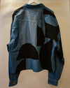 A blue denim jacket with large black abstract patchwork panels on the back, hanging on a white hanger against a plain beige wall. Caroline Dickinson Shirt | Fodder