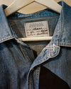 A close-up of a blue denim shirt jacket hanging on a white hanger, showing the collar, front button placket, and an interior neck label that reads Caroline Dickinson. Caroline Dickinson Shirt | Fodder