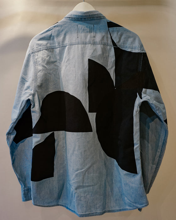 A light blue denim shirt jacket shown from the back, featuring large abstract black patchwork shapes over a light blue denim base, hanging on a white hanger against a plain beige wall. Caroline Dickinson Shirt | Fodder