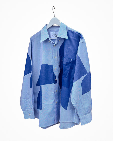 A long-sleeve button-up shirt with a geometric patchwork pattern in shades of blue, hanging on a white hanger against a plain white background. Caroline Dickinson Shirt | Fodder