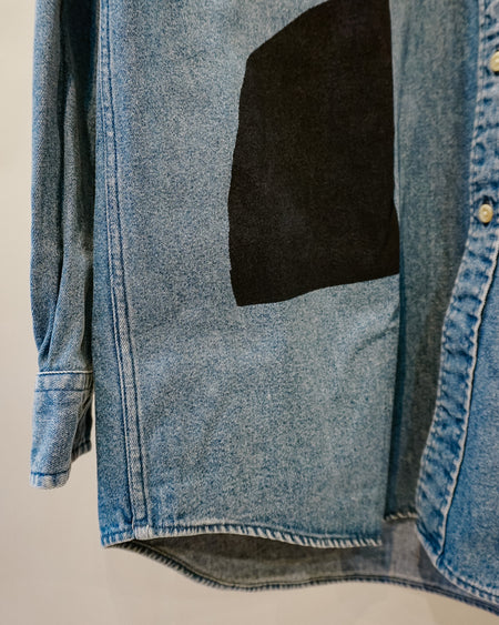 A close-up of a light blue denim button-up shirt with a large black fabric patch on the front, shown hanging against a plain light background. Caroline Dickinson Shirt | Fodder