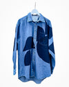 A blue denim button-up shirt with abstract dark blue patchwork panels, hanging on a white hanger against a plain white background. Caroline Dickinson Shirt | Fodder