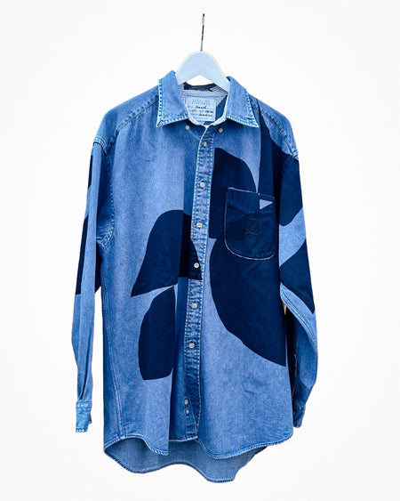 A blue denim button-up shirt with abstract dark blue patchwork panels, hanging on a white hanger against a plain white background. Caroline Dickinson Shirt | Fodder