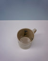 Beige mug with black design on a white surface and light blue background. Tulip Mug | Nerikomi | Fern Taylor | Shop Fodder