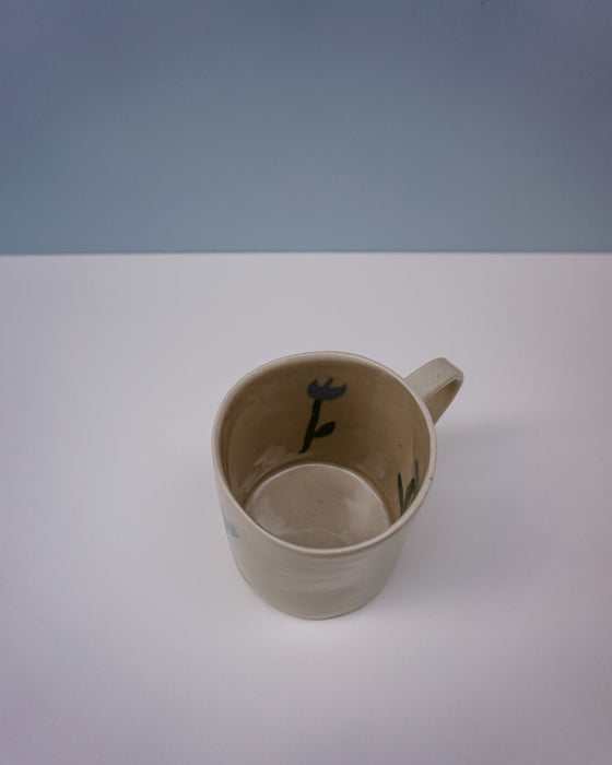 Beige mug with black design on a white surface and light blue background. Tulip Mug | Nerikomi | Fern Taylor | Shop Fodder
