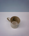 Beige mug with cartoon character on a white surface and blue background. Tulip Mug | Nerikomi | Fern Taylor | Shop Fodder