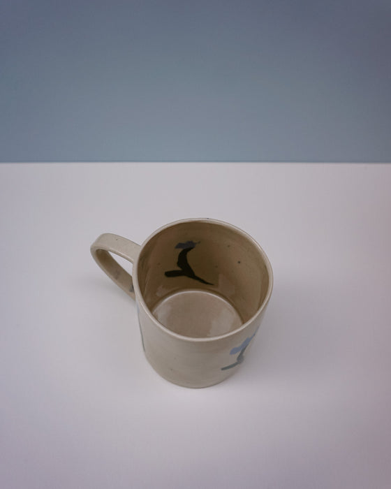 Beige mug with cartoon character on a white surface and blue background. Tulip Mug | Nerikomi | Fern Taylor | Shop Fodder