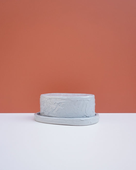 A small round gray textured planter pot with a matching saucer, set against a split background of white and terracotta orange. Brutes Ceramics | Hope Collection | Shop Fodder