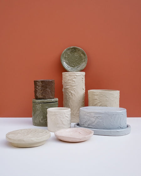 A collection of textured ceramic planters and shallow dishes in earthy tones, including beige, gray, green, and pink, arranged on a white surface against a terracotta background. Brutes Ceramics | Hope Collection | Shop Fodder
