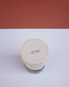 A handmade ceramic tin with a round, flat lid, featuring a stamped 'brutes' logo on the top. The tin has a textured, hand-painted beige finish with a dark brown base, set against a split white and terracotta background. Brutes Ceramics | Hope Collection | Shop Fodder