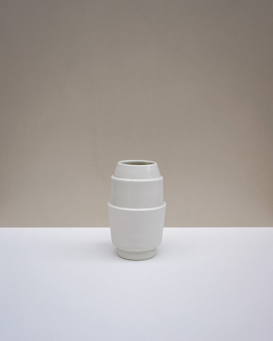 A minimalist white ceramic vase with a stacked, geometric barrel-like design, set against a neutral beige and white studio background. Emma Johnson Ceramics | Fodder | Hope Collection