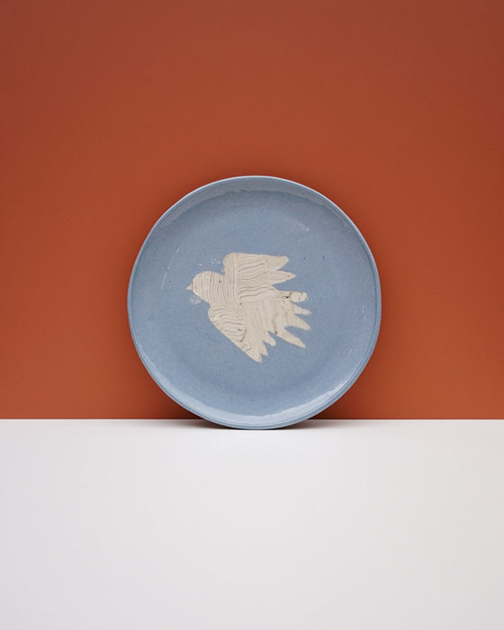 A round light blue ceramic plate with a textured off-white bird design in the center, set against a split orange and white background. Fern Taylor Ceramics | Hope Collection | Shop Fodder