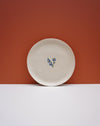 A round off-white ceramic plate with a small blue and green floral motif in the center, set against a split orange and white background. Fern Taylor Ceramics | Hope Collection | Shop Fodder