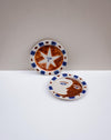 Two decorative ceramic plates with a hand-painted style design, featuring a brown and white face and a brown star with blue accents, displayed on a two-tone white and gray surface. Hoodmor Studio | Fodder | Hope Collection