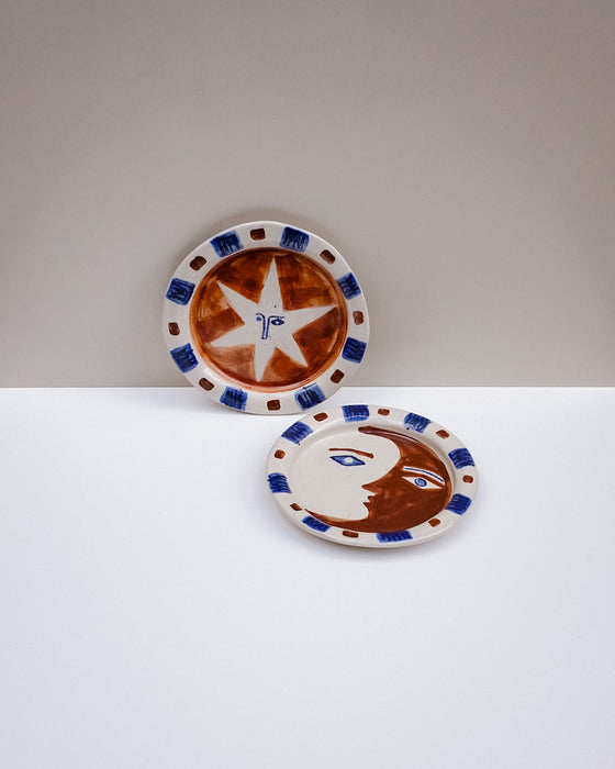 Two decorative ceramic plates with a hand-painted style design, featuring a brown and white face and a brown star with blue accents, displayed on a two-tone white and gray surface. Hoodmor Studio | Fodder | Hope Collection