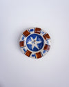 A small round ceramic dish with a hand-painted design featuring a blue and white star in the center and brown and white geometric border patterns, set against a plain light gray background. Hoodmor Studio | Fodder | Hope Collection