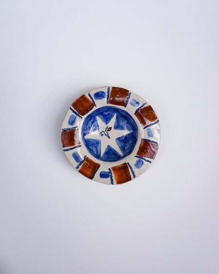 A small round ceramic dish with a hand-painted design featuring a blue and white star in the center and brown and white geometric border patterns, set against a plain light gray background. Hoodmor Studio | Fodder | Hope Collection