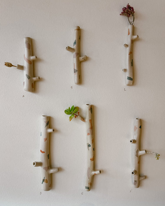 Six hand-painted ceramic tree branch wall hooks in natural beige clay with colorful speckles, arranged on a light beige wall with small decorative plants. Jode Pankhurst Ceramics. Fodder