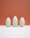 Three small matte ceramic vases in a speckled off-white finish, arranged in a row on a white surface against a solid terracotta background. KJA Studio Ceramics | Hope Collection | Shop Fodder