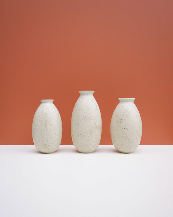 Three small matte ceramic vases in a speckled off-white finish, arranged in a row on a white surface against a solid terracotta background. KJA Studio Ceramics | Hope Collection | Shop Fodder