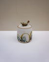 A small handmade ceramic lidded jar with a bird-shaped lid topper, featuring a white glaze with gold, green, and blue accents, set against a neutral beige and white background.  Liz Vidal Ceramics | Fodder | Hope Collection