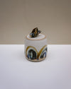 A small handmade ceramic lidded container with a lid topped by a small seashell figurine. The ceramic body is off-white with a glazed finish and features abstract painted patterns in blue, gold, and brown.  Liz Vidal Ceramics | Fodder | Hope Collection