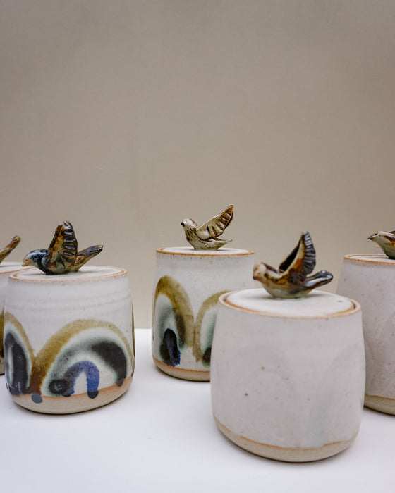 A set of handmade ceramic lidded jars with flying bird toppers, featuring a white glaze and hand-painted abstract arches in earthy tones.  Liz Vidal Ceramics | Fodder | Hope Collection