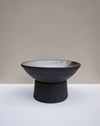 A dark charcoal ceramic pedestal bowl with a light gray glazed interior, set against a neutral beige and white studio background. Scree Ceramics | Fodder | Hope Collection