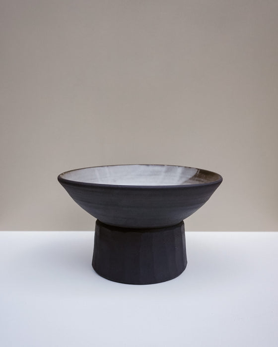 A dark charcoal ceramic pedestal bowl with a light gray glazed interior, set against a neutral beige and white studio background. Scree Ceramics | Fodder | Hope Collection
