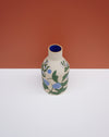 A small ceramic bottle with a beige body, decorated with green leaves and blue berries, featuring a deep blue interior, set against a split white and terracotta background. Sophie Alda | Hope Collection | Shop Fodder