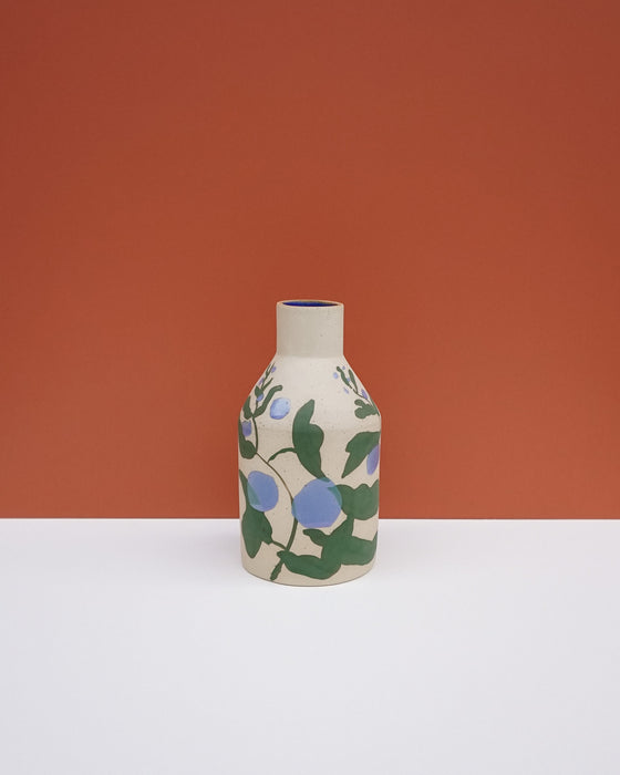 A small ceramic vase with a cream-colored body, decorated with a green and blue floral pattern, standing on a white surface against a terracotta orange background. Sophie Alda | Hope Collection | Shop Fodder