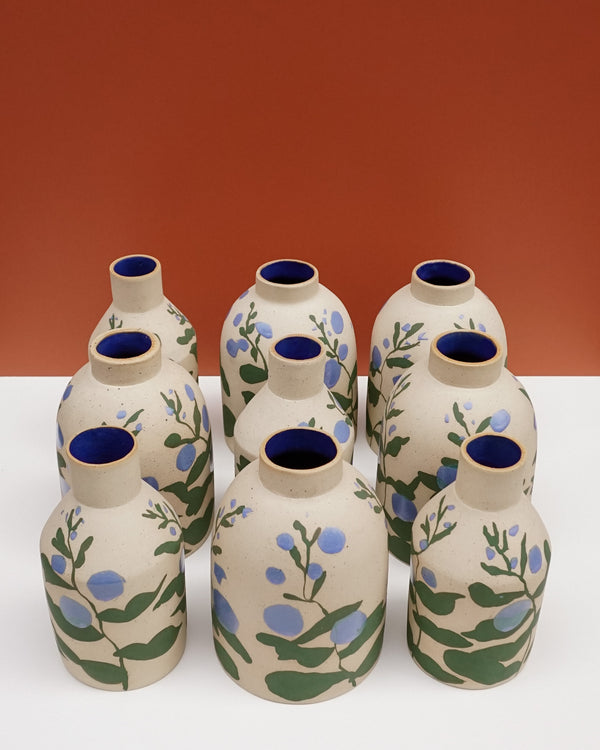 Eight hand-painted ceramic bottles with beige bodies, blue interiors, and green and blue floral motifs, arranged on a white surface against a terracotta background.  Sophie Alda | Hope Collection | Shop Fodder