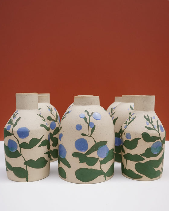 Three handmade ceramic vases with a beige stoneware finish, decorated with a hand-painted pattern of green leaves and blue berries, set against a solid terracotta background. Sophie Alda | Hope Collection | Shop Fodder