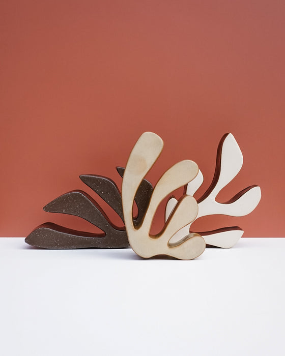 Three abstract leaf-shaped wall art sculptures in beige, brown, and white, arranged in a fan shape against a terracotta background and white surface. Studio ERS Ceramics | Hope Collection | Shop Fodder
