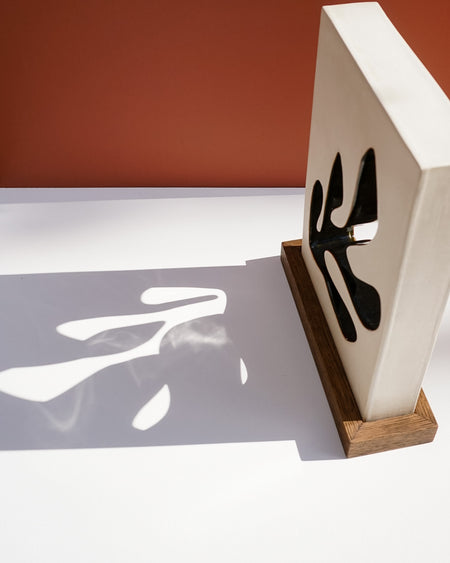 A modern decorative wall art piece featuring a white rectangular panel with black abstract cutout shapes, mounted on a wooden base and casting a bold shadow on a white surface against a terracotta background. Studio ERS Ceramics | Hope Collection | Shop Fodder
