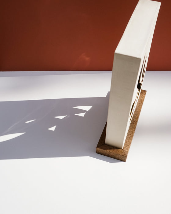 A modern, minimalist wall clock with a white rectangular face and a natural wood base, shown standing upright on a white surface with dramatic shadows against a terracotta background. Studio ERS Ceramics | Hope Collection | Shop Fodder