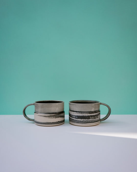 Two ceramic mugs with striped design on a light blue surface and teal background.  Janne Meijer Ceramics | Shop Fodder