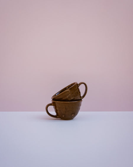 Two stacked brown ceramic cups with a textured, basket-like exterior and looped handles, set against a minimalist two-tone background of soft pink and white.